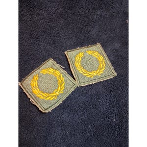 May include: Two square-shaped embroidered patches featuring an olive green fabric base with a bright yellow laurel wreath design stitched in the center. The edges of the patches are finished with a matching olive green thread border.