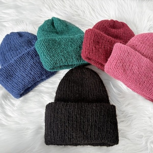 Peut inclure: Plusieurs bonnets tricotés de couleurs variées : bleu, vert, bordeaux, rose et noir. Ils sont disposés sur une surface blanche et duveteuse. Chaque bonnet a un revers plié.