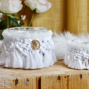 May include: Two white macrame candle holders with a gold coin detail. The holders have a fringe and faux fur trim. They are displayed on a wooden surface with white feathers and flowers in the background.