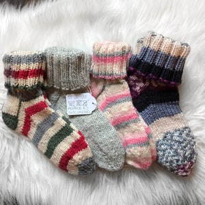 May include: Four pairs of colorful, hand-knitted socks with folded cuffs. The socks feature various patterns and color combinations, including stripes and geometric designs. A tag with text is attached to one sock.
