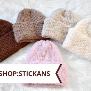 Peut inclure: Cinq bonnets tricotés dans des tons marron foncé, marron moyen, beige, crème et rose clair. Chaque bonnet présente un revers plié et une maille texturée duveteuse. Une bannière blanche en bas affiche le texte NEW SHOP:STICKANS.