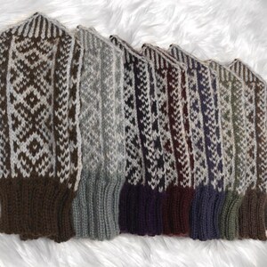 May include: Seven knitted mittens in a range of colours, including brown, grey, burgundy, and blue. Each mitten has a geometric white pattern and a contrasting cuff. The mittens are displayed on a white, fluffy surface.