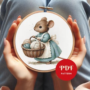 May include: Cross-stitch pattern of a mouse in a blue dress and white apron carrying a basket of yarn. The mouse is brown with a long tail and whiskers. The design is in a wooden embroidery hoop. The text "PDF PATTERN" is in a red circle.