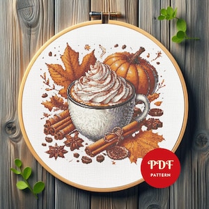May include: Cross-stitch pattern featuring a fall theme. A white mug with whipped cream and cinnamon sits next to a pumpkin, fall leaves, and cinnamon sticks. The design is set in a wooden embroidery hoop. The text "PDF PATTERN" is in a red circle.