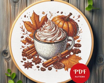 May include: Cross-stitch pattern featuring a fall theme. A white mug with whipped cream and cinnamon sits next to a pumpkin, fall leaves, and cinnamon sticks. The design is set in a wooden embroidery hoop. The text "PDF PATTERN" is in a red circle.