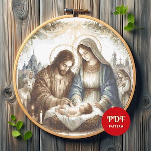 May include: A circular embroidery hoop containing a cross-stitch pattern of the Nativity scene. The image depicts a bearded figure and a veiled figure looking down at an infant in a manger, with a city silhouette in the background. A red PDF PATTERN label is visible.