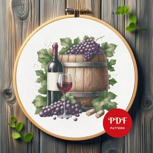 Puede incluir: Patrón de punto de cruz con temática de vino. El diseño presenta una botella de vino, una copa de vino tinto, una barrica de madera y uvas. El diseño está en un bastidor de bordado de madera. Un círculo rojo con el texto "PDF PATTERN" está en la esquina inferior derecha.