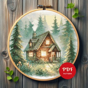 May include: A cross-stitch pattern of a cozy cabin nestled in a forest, framed in a wooden embroidery hoop. The cabin has a stone chimney and lit windows, surrounded by evergreen trees and a misty atmosphere. The text "PDF PATTERN" is in a red circle.