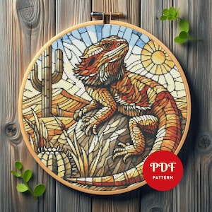 Könnte beinhalten: Ein Kreuzstichmuster eines Bartagamen in einer Wüstenlandschaft, eingerahmt in einem hölzernen Stickrahmen. Das Design zeigt eine Eidechse in Orange-, Rot- und Gelbtönen, einen Kaktus und eine Sonne. Der Text "PDF PATTERN" befindet sich in einem roten Kreis.