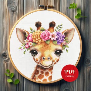 Puede incluir: Patrón de punto de cruz de una jirafa con una corona de flores, exhibido en un bastidor de bordado de madera. La jirafa tiene manchas marrones y ojos grandes. La corona floral incluye flores rosas, amarillas y moradas. Un círculo rojo con "PDF PATTERN" está en la parte inferior derecha.