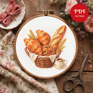 May include: A cross-stitch pattern in a wooden embroidery hoop featuring a woven basket filled with golden-brown croissants, a baguette, and wheat stalks, accompanied by a small cup of coffee. A red circular badge displays the text PDF PATTERN.