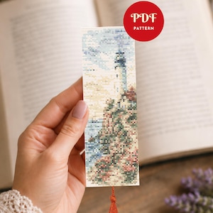 以下が含まれることがあります： 青い空と海を背景に、崖の上の灯台を描いたクロスステッチのブックマーク。 ブックマークにはタッセルが付いており、赤い円に「PDF PATTERN」という文字が書かれています。