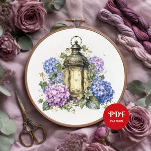 May include: A cross stitch pattern in a wooden embroidery hoop featuring a vintage lantern surrounded by blue and purple hydrangea flowers and green leaves. The design is set against a white fabric background with a red circular PDF pattern label.