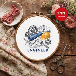 May include: A cross-stitch pattern in a wooden embroidery hoop featuring engineering icons including gears, a bridge, a blue blueprint, a yellow hard hat, a compass, a wrench, and a bearing, with the word ENGINEER printed in dark blue text.