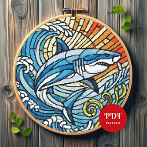 Peut inclure: Motif de point de croix d'un requin dans les vagues de l'océan, encadré dans un cercle à broder en bois. Le motif utilise des nuances de bleu, blanc, jaune et orange, avec un effet de vitrail. Un cercle rouge avec le texte "PDF PATTERN" est en bas à droite.