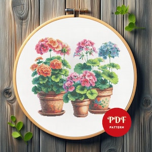May include: A cross-stitch pattern featuring colorful geraniums in terracotta pots. The embroidery is set in a wooden hoop, with a red circle that says "PDF PATTERN". The geraniums have pink, orange, and blue flowers.