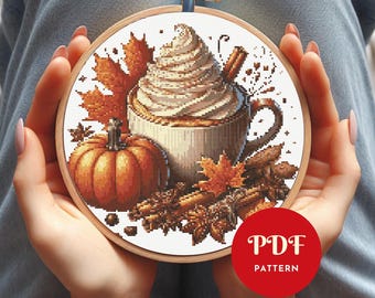 May include: A cross-stitch pattern in a wooden hoop features a fall theme. The design includes a pumpkin, a coffee cup with whipped cream, cinnamon sticks, and autumn leaves. The text "PDF PATTERN" is in a red circle.