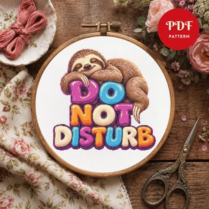 Puede incluir: Un patrón de punto de cruz en un bastidor de madera que muestra un perezoso marrón durmiendo sobre letras coloridas estilo burbuja que forman el texto DO NOT DISTURB. Las letras son rosas, azules, naranjas y crema con contornos oscuros.