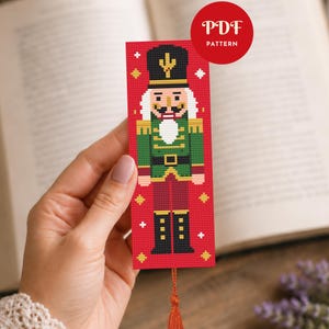 May include: A rectangular cross-stitch bookmark featuring a nutcracker design on a red background with small white and yellow star accents. The nutcracker wears a black hat, green jacket, and dark red trousers. A red tassel hangs from the bottom.