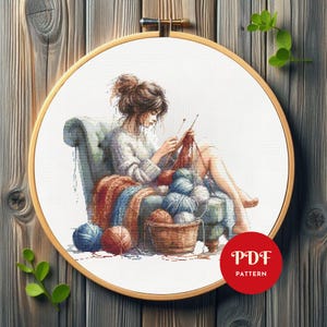 Può includere: Uno schema di punto croce che presenta una donna seduta su una sedia, a maglia con gomitoli di lana e ferri. La donna indossa un maglione bianco e ha i capelli castani. La sedia è di colore blu chiaro e ha una coperta marrone drappeggiata sopra di essa. Lo sfondo è una tela bianca.