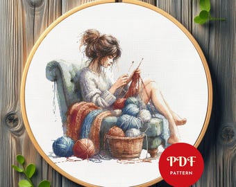 Può includere: Uno schema di punto croce che presenta una donna seduta su una sedia, a maglia con gomitoli di lana e ferri. La donna indossa un maglione bianco e ha i capelli castani. La sedia è di colore blu chiaro e ha una coperta marrone drappeggiata sopra di essa. Lo sfondo è una tela bianca.