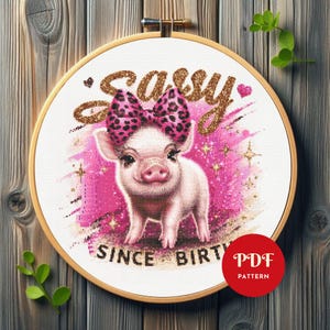 Może przedstawiać: Haft krzyżykowy przedstawiający prosię w różowej kokardzie w panterkę, ze słowem "Sassy" w złotych, brokatowych literach. Tekst "SINCE BIRTH" znajduje się pod świnią. Projekt umieszczono w drewnianej tamborku.