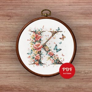 May include: Cross-stitch embroidery of the letter "K" adorned with pink and yellow flowers and blue butterflies, set in a wooden embroidery hoop. A red circle with "PDF PATTERN" text is visible.