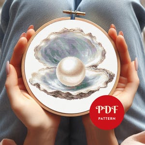Può includere: Ricamo a punto croce di una perla all'interno di una conchiglia aperta, in un telaio di legno. Il guscio è nei toni del blu, grigio e bianco, con una grande perla bianca e rotonda al centro. Un cerchio rosso con il testo "PDF PATTERN" è in basso a destra.
