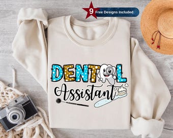 Op de afbeelding: Een wit sweatshirt met de tekst "DENTAL ASSISTANT" in blauwe, gouden en witte letters met stippen. Een cartoon-tand met een vergrootglas bevindt zich boven de tekst. Een tandenborstel en een tandheelkundig instrument bevinden zich onder de tekst.