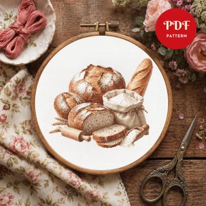 May include: A cross stitch pattern in a wooden embroidery hoop featuring a collection of baked bread, a small sack of flour, a wooden rolling pin, and a scoop of flour. The design is printed on white fabric with a red PDF PATTERN label.