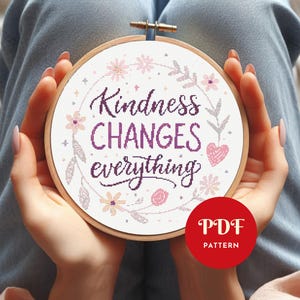 May include: A cross-stitch embroidery hoop featuring the phrase "Kindness Changes Everything" in purple script. The design incorporates floral and heart motifs in pink and lavender. A red circle with "PDF PATTERN" is at the bottom. The background is white.