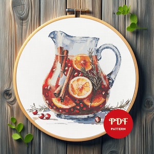 Könnte beinhalten: Kreuzstichmuster eines Glaskruges gefüllt mit einem roten Getränk, Orangenscheiben, Zimtstangen und Rosmarin. Das Design befindet sich in einem hölzernen Stickrahmen. Ein roter Kreis mit "PDF PATTERN" befindet sich unten rechts.