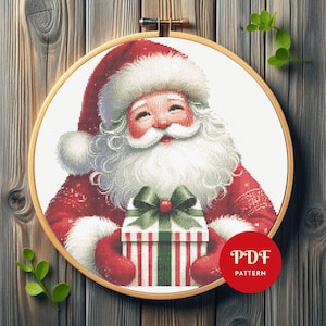 May include: Cross-stitch pattern of Santa Claus holding a wrapped gift. Santa is wearing a red suit and hat with white trim, and has a long white beard. The gift is wrapped in white paper with red and green stripes, and a green bow. The pattern is in a wooden embroidery hoop.