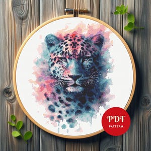 May include: A cross-stitch pattern of a leopard's face, featuring a watercolor effect with blue, pink, and purple hues. The design is set within a wooden embroidery hoop. A red circle with the text "PDF PATTERN" is in the lower right corner.