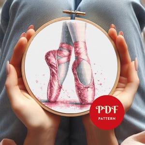 May include: Cross-stitch embroidery hoop featuring a watercolor design of pink ballet pointe shoes. The image includes a red circle with the text "PDF PATTERN". The design is set against a white background with pink accents.