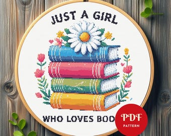 May include: Cross-stitch embroidery in a wooden hoop. The design features a stack of colorful books with a daisy on top, surrounded by flowers. Text reads "JUST A GIRL WHO LOVES BOOKS." A red circle with "PDF PATTERN" is in the lower right.