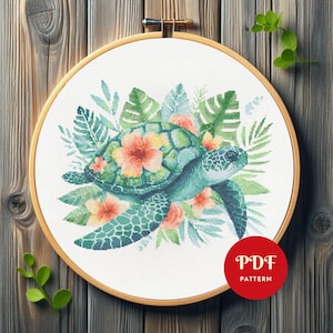 May include: A cross-stitch embroidery pattern in a wooden hoop featuring a sea turtle surrounded by tropical green leaves and orange flowers. A red circular badge in the bottom right corner contains the white text PDF PATTERN.