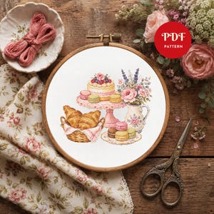 May include: A cross-stitch pattern in a wooden embroidery hoop featuring a cake on a pink stand, a basket of croissants, colorful macarons, and a floral pitcher. The design includes the text PDF PATTERN in a red circle on the upper right side.