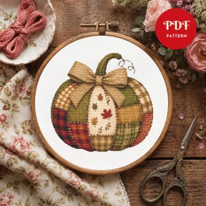 Könnte beinhalten: Ein Kreuzstichmuster eines Kürbisses in einem hölzernen Stickrahmen. Der Kürbis zeigt ein Patchwork-Design mit rot-schwarzen Karomustern, grünen und beigen Abschnitten, einem zentralen Feld mit Herbstblättern und einer großen beigen Schleife.
