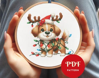 May include: A cross-stitch embroidery hoop featuring a cute puppy wearing a Santa hat with reindeer antlers and wrapped in Christmas lights. The puppy is brown and white, with a smiling expression. The hoop is held by hands.