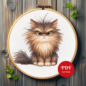 May include: A cross-stitch pattern of a grumpy-looking cat with brown and tan fur, set in a wooden embroidery hoop. The cat has an angry expression with furrowed brows. A red circle with "PDF PATTERN" is in the lower right.
