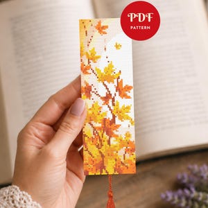 May include: A rectangular cross-stitch bookmark featuring a pattern of autumn maple leaves in shades of yellow, orange, and brown against a white background, finished with a small orange tassel at the bottom and a red circular PDF PATTERN label.