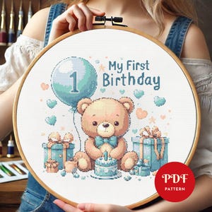以下が含まれることがあります： かわいいテディベアが1本のろうそくのついた誕生日ケーキを持っているクロスステッチパターン。くまはプレゼントと数字1のついた青い風船に囲まれています。「My First Birthday」という文字がくまの上に書かれています。パターンはPDFダウンロードで入手できます。