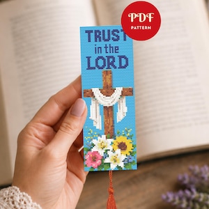 Könnte beinhalten: Ein rechteckiges Kreuzstich-Lesezeichen mit hellblauem Hintergrund, dem dunkelblauen Text TRUST in the LORD über einem braunen Holzkreuz mit weißem Tuch, verziert mit weißen, gelben und rosa Blumen an der Basis und einer roten Quaste.