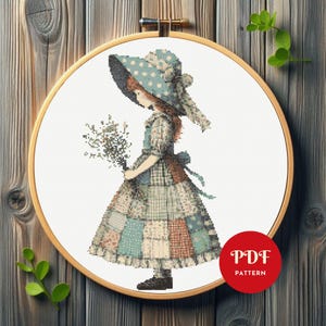 May include: A cross stitch pattern featuring a girl in a blue and white polka dot hat and a patchwork dress holding a bouquet of flowers. The pattern is in a hoop and the text "PDF PATTERN" is visible in the lower right corner.