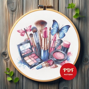 Op de afbeelding: Kruissteekborduurwerk van make-upproducten, waaronder lippenstiften, kwasten en een oogschaduwpalet. Het ontwerp toont roze en blauwe vlinders en is geplaatst in een houten borduurring. De tekst "PDF PATTERN" staat in een rode cirkel.