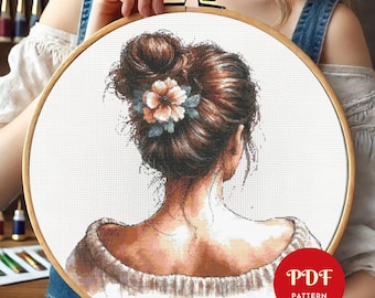 Può includere: Schema di punto croce di una donna con capelli castani raccolti in uno chignon, che indossa un maglione bianco. La donna è di spalle e ha un fiore tra i capelli. Lo schema è in un cerchio e il testo "PDF PATTERN" è visibile in basso a destra.