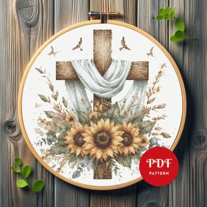 Op de afbeelding: Een kruissteekpatroon met een houten kruis, bedekt met witte stof, versierd met zonnebloemen en bladeren. Het ontwerp is geplaatst in een houten borduurring. De afbeelding bevat een rode cirkel met de tekst "PDF PATTERN".