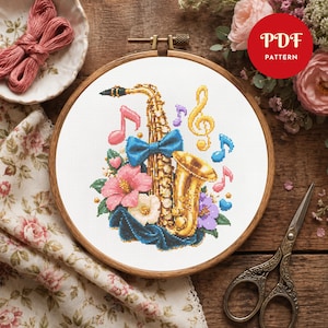 Peut inclure: Un modèle de point de croix dans un tambour à broder en bois représentant un saxophone doré orné d'un nœud bleu, de fleurs d'hibiscus roses et blanches, et de notes de musique colorées dont une clé de sol. Le texte PDF PATTERN est visible.