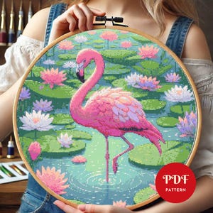 Puede incluir: Un bastidor de madera circular que contiene un patrón de punto de cruz de un flamenco rosa brillante de pie en un estanque lleno de nenúfares verdes y flores de loto rosas y blancas. Una insignia roja con el texto blanco PDF PATTERN.